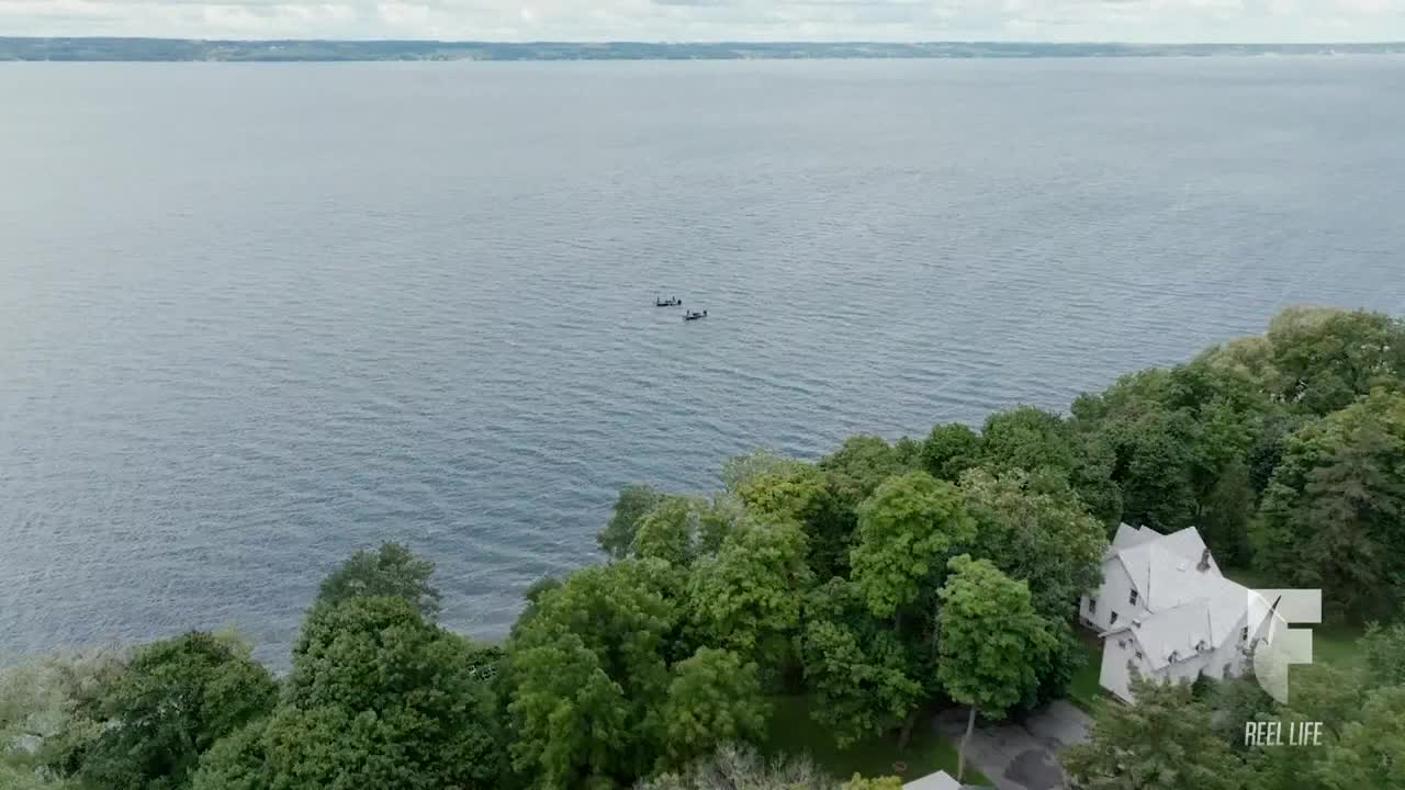 Two small boats are gliding across the water, far from the shoreline. Lush green trees frame a large white house nestled on the edge of the lake.
