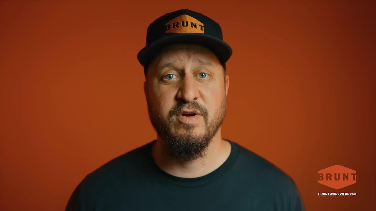 A man with a beard and a BRUNT hat is speaking directly to the camera, his mouth open. The background is a solid orange color, and the BRUNT logo is visible in the lower right corner.
