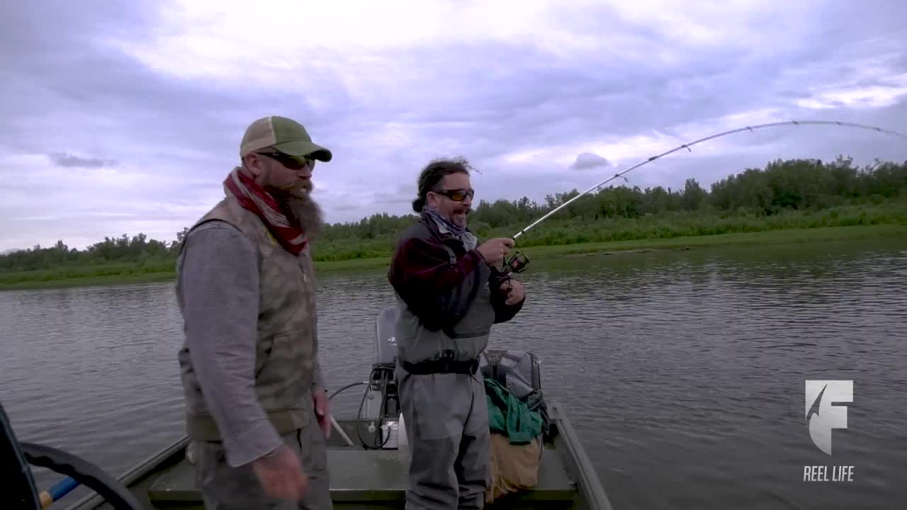 A man in waders is reeling in a fishing line, the rod bent nearly double. His companion watches from the boat as they fish the cloudy waters.
