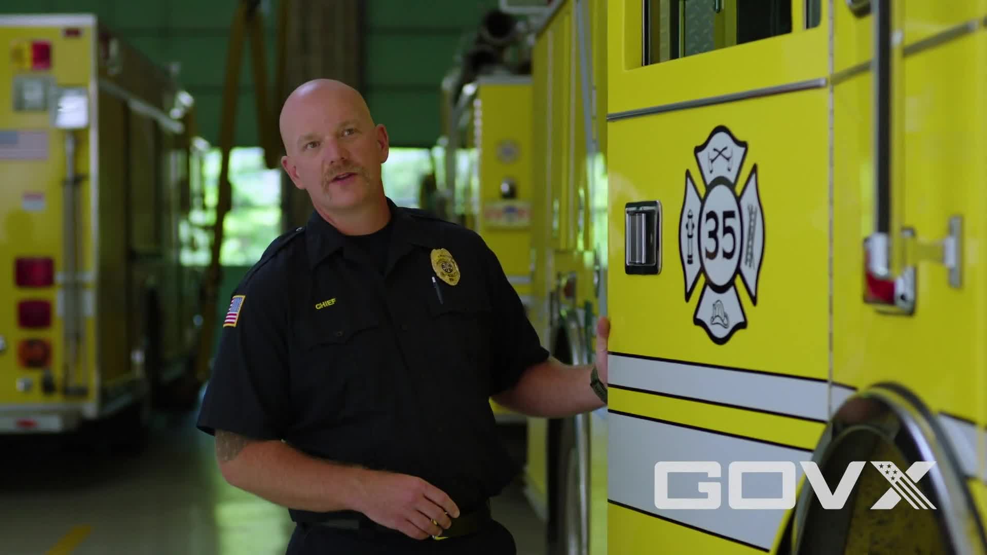 A firefighter in uniform stands next to a bright yellow fire truck, gesturing toward the camera. The number "35" is displayed prominently on the side of the vehicle.
