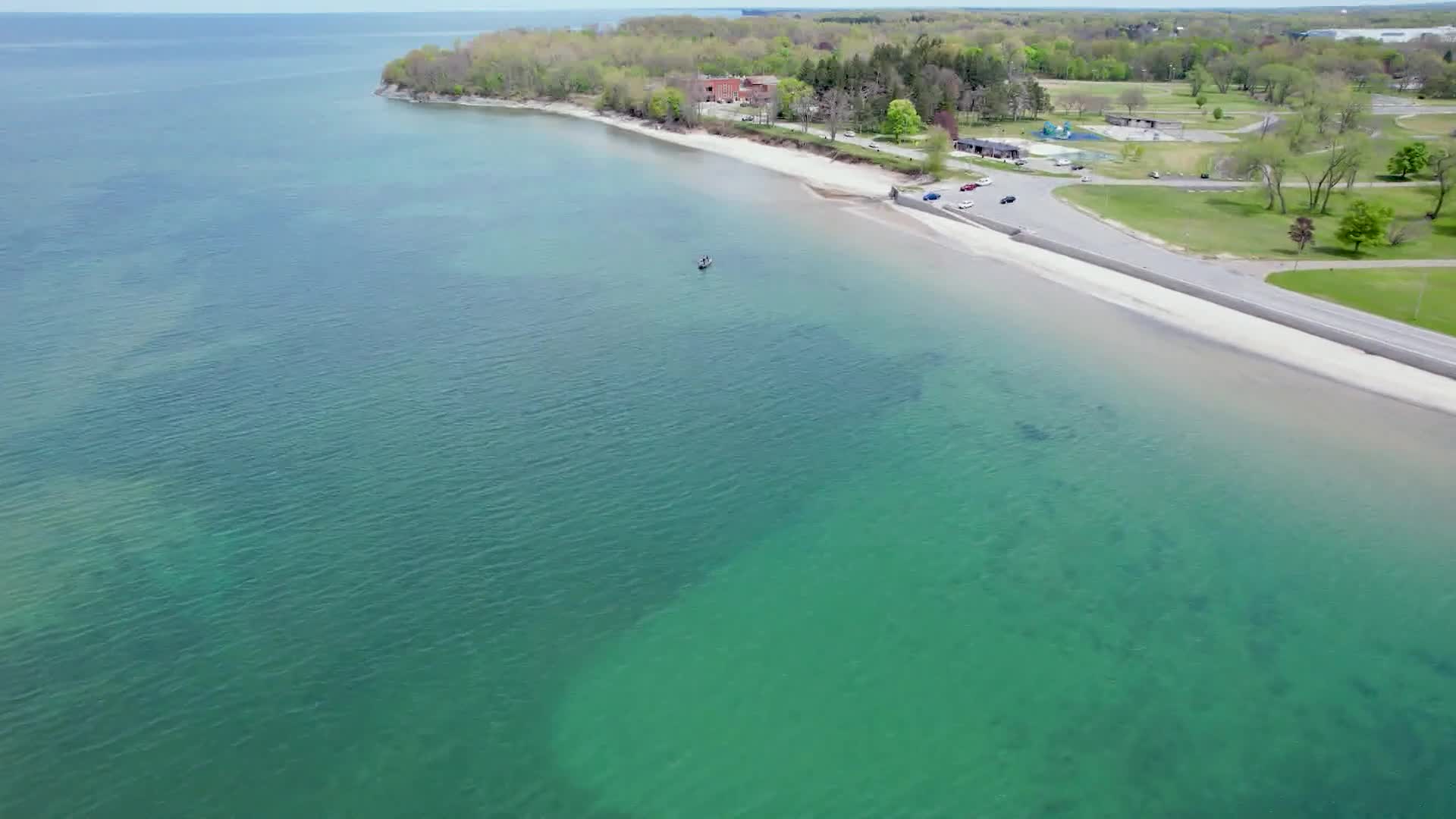 A lone figure paddles a small craft on the clear turquoise water near the shoreline. The beach curves along the coast, with a road and parking area just beyond the sand.
