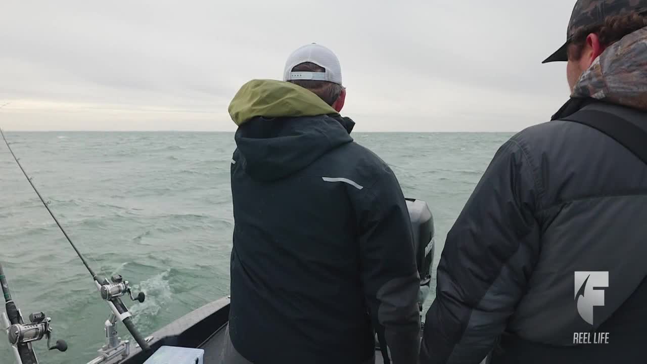 Two anglers in dark jackets scan the choppy water from a boat, rods at the ready. The overcast sky and gray waves suggest a challenging day on the water.
