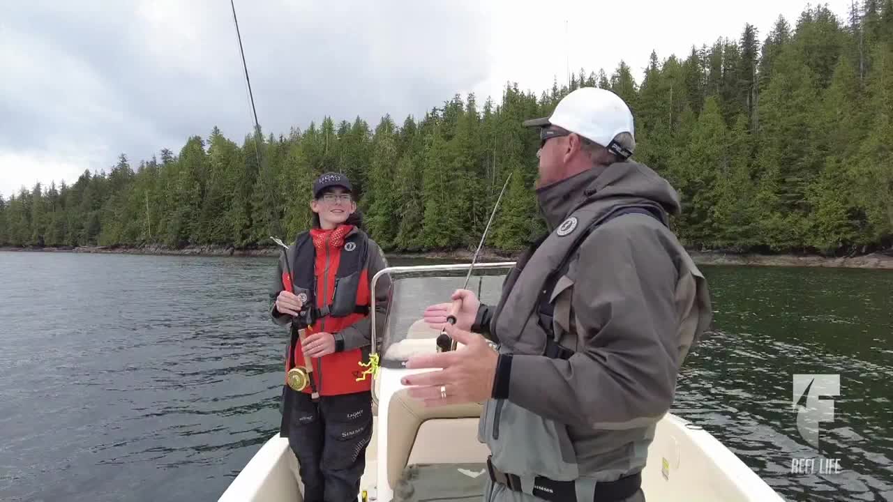 A man in a white cap gestures with his hands, explaining something to a young angler holding a fishing rod. They're in a small boat on a calm body of water, with a dense forest lining the shore.
