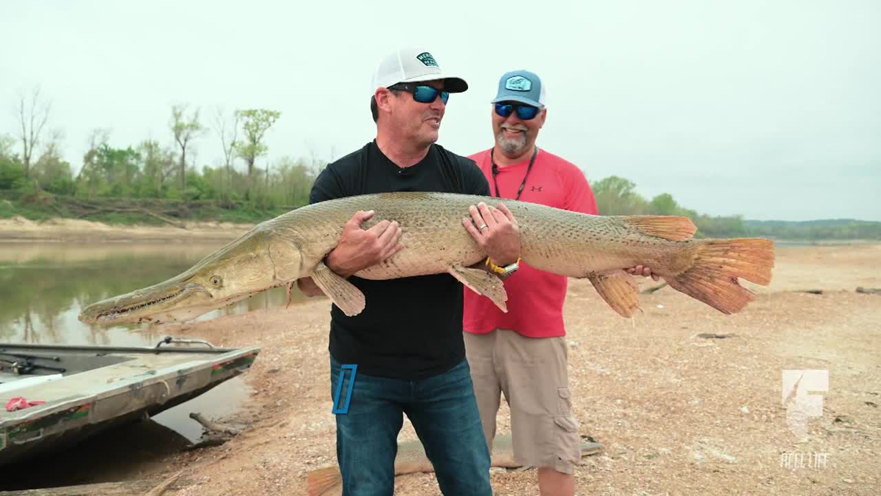 Two men stand on a muddy bank, grinning as they hold up a massive alligator gar. The fish's long snout and orange tail are clearly visible as they pose for the camera.
