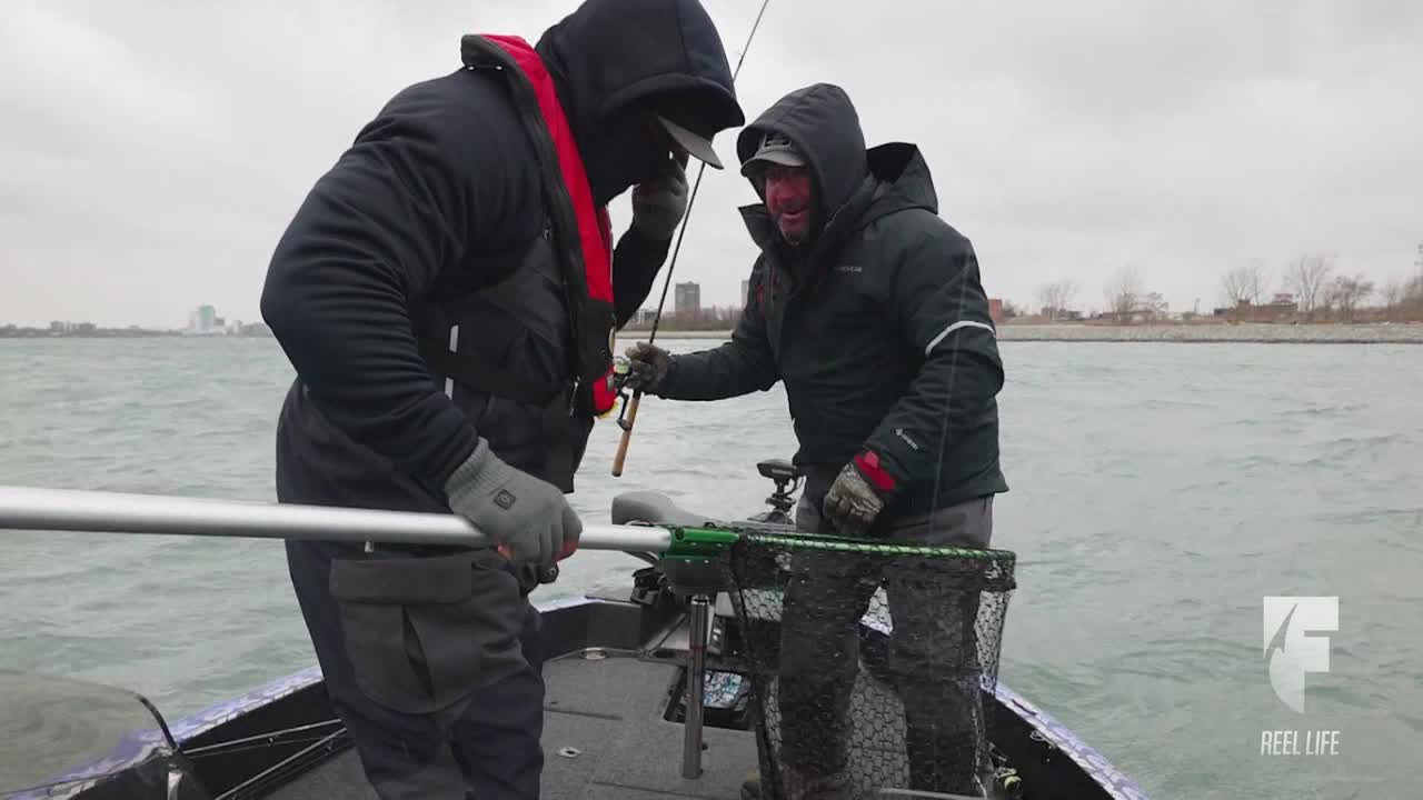 Two anglers, bundled up against the cold, work together on a boat in the United States, likely on the World Fishing Network's "Reel Life." One man carefully guides a fishing rod while the other holds a net, ready to scoop up a catch.

