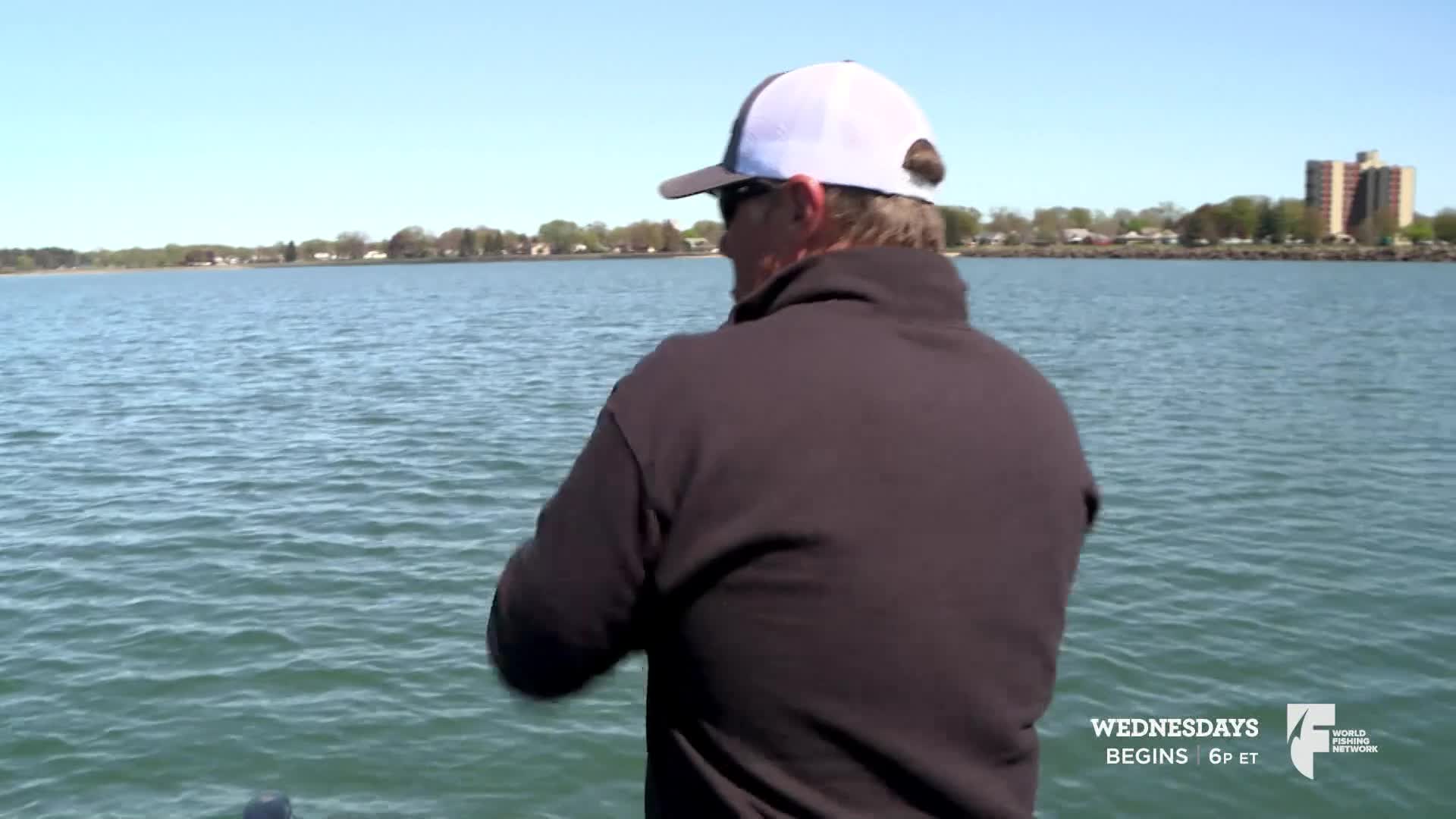 A man in a cap and sunglasses is facing away from me, casting a line into the water. The calm lake reflects the clear sky as he fishes, likely on a show from the World Fishing Network.
