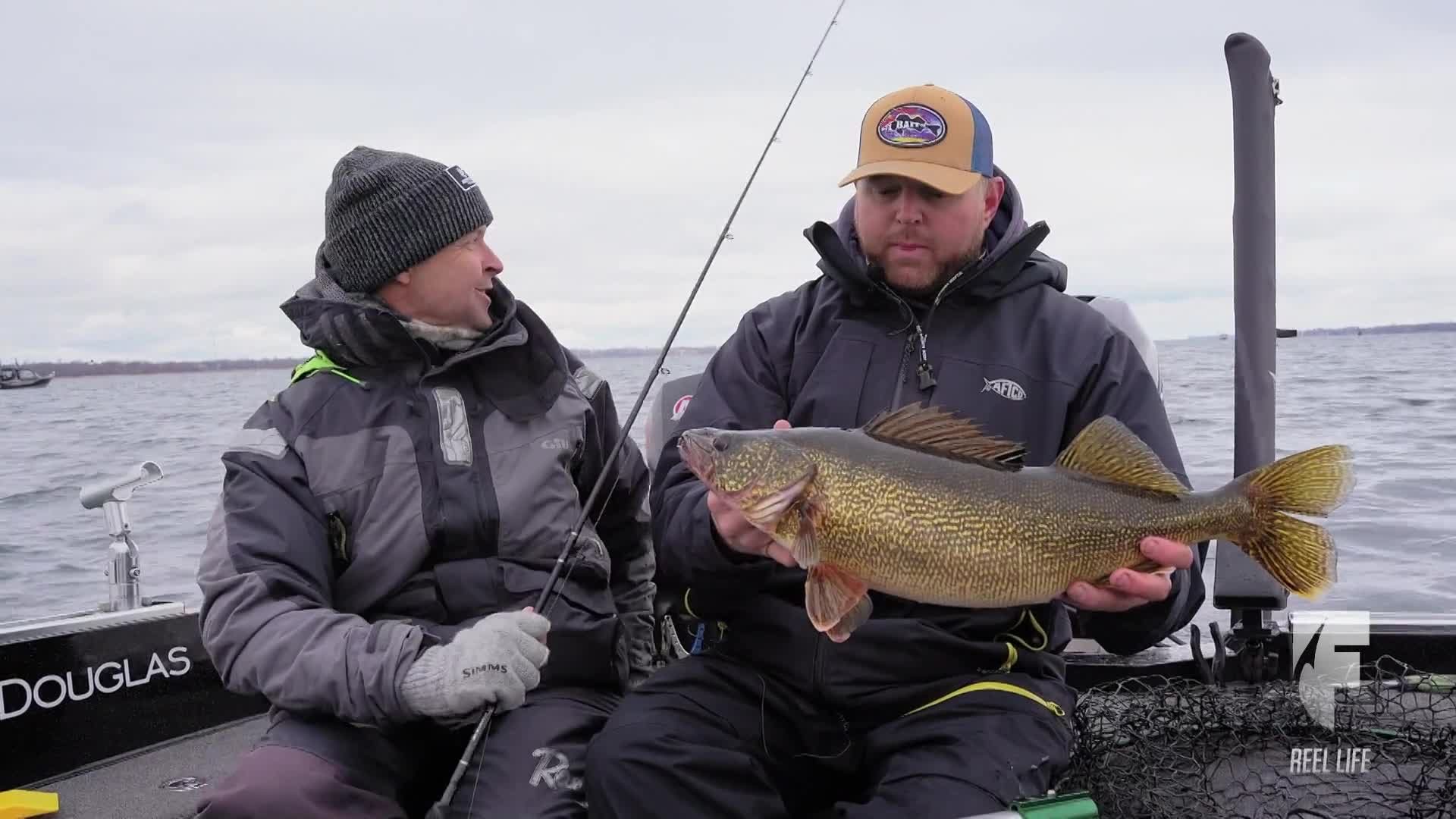 A fisherman, wearing a ball cap, holds up a large walleye, its golden scales catching the light. The other man, in a knit cap, looks on, smiling, as the boat gently bobs on the water.
