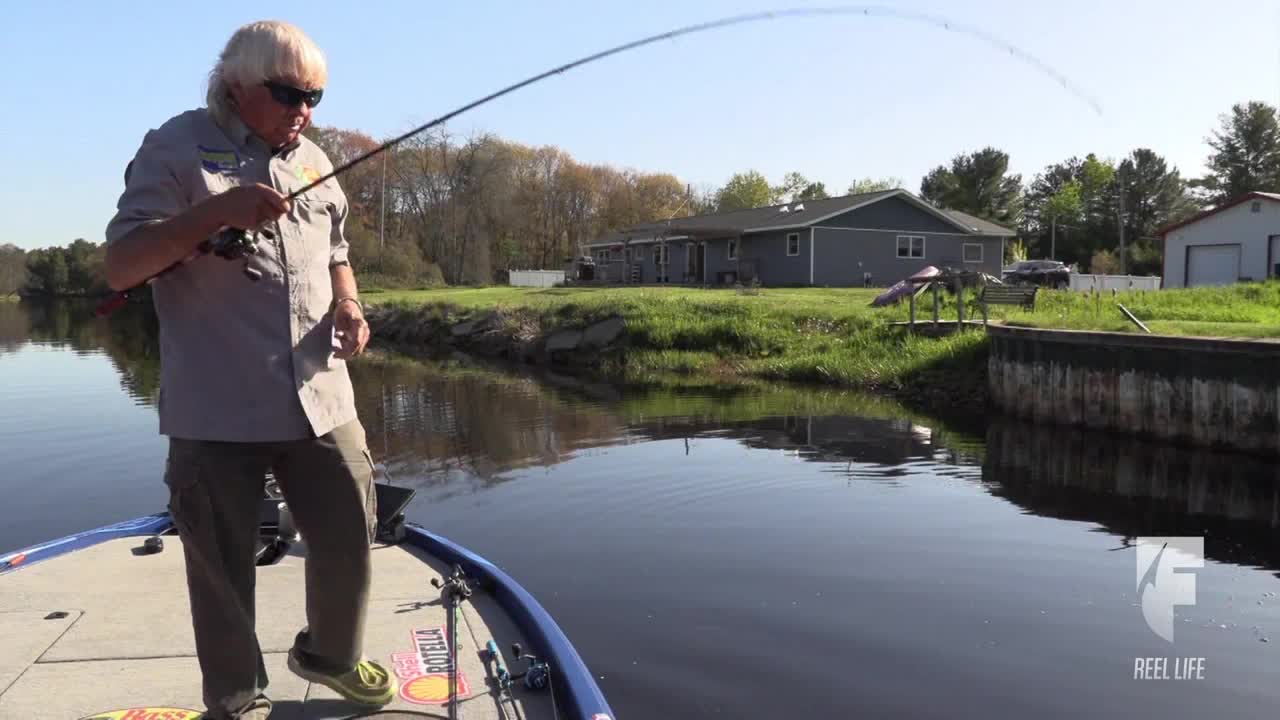 The man on the boat is holding a fishing rod that's bent nearly in half, the line taut. He's fishing in the calm water, with a house and a dock in the background.
