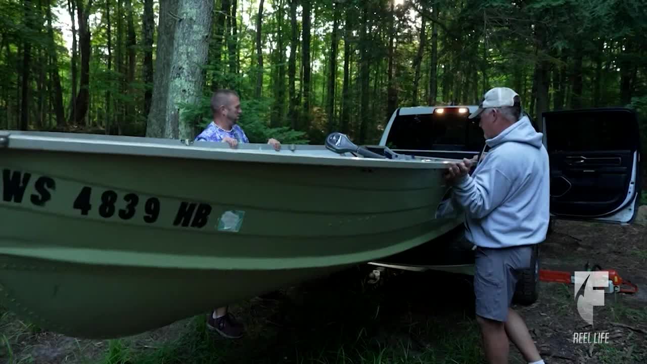 Two men are lifting an aluminum boat from the back of a truck. They're in a wooded area, likely preparing for a day of fishing.
