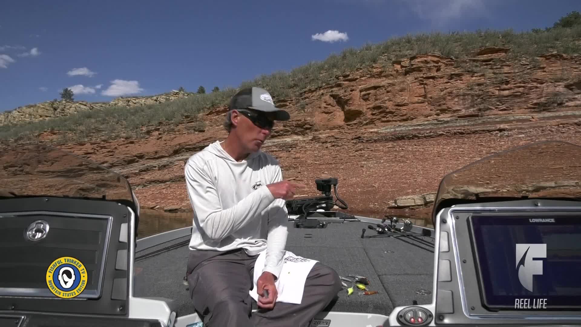 A man in a fishing boat gestures towards a device, while a document sits in his lap. Behind him, a rocky, arid landscape rises under a bright blue sky.
