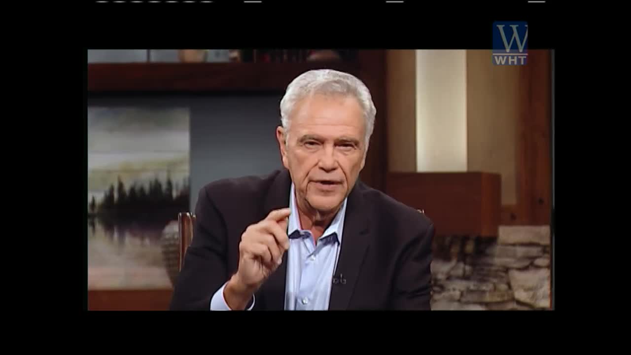 The man in the suit gestures with his index finger as he speaks, likely on World Harvest TV. He's sitting in a studio, a landscape painting visible behind him.
