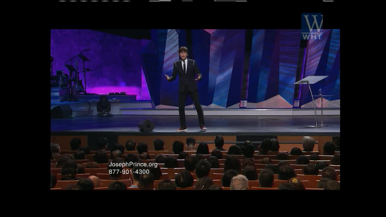 A man in a suit gestures with both hands on a stage, likely during a World Harvest TV broadcast. The audience is seated, their backs to me, focused on the man.
