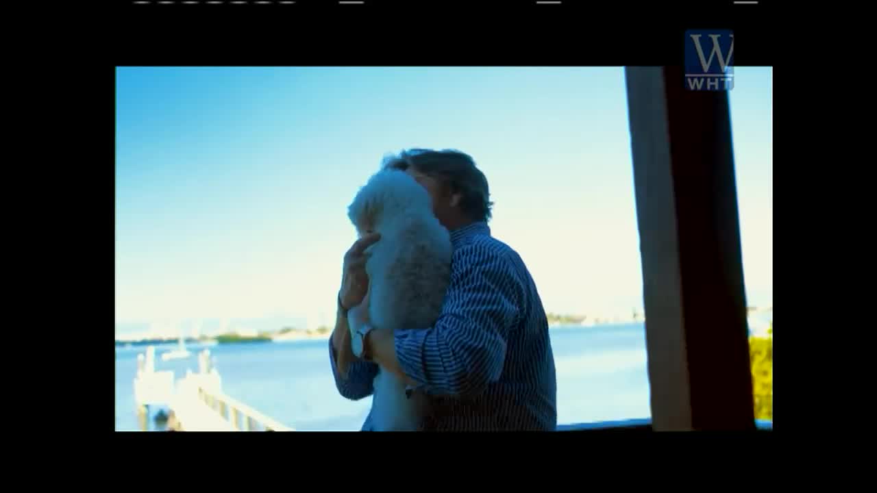 A man in a blue striped shirt is holding a fluffy white dog close. The scene is framed by a waterfront view, likely from a veranda, as seen on World Harvest TV.
