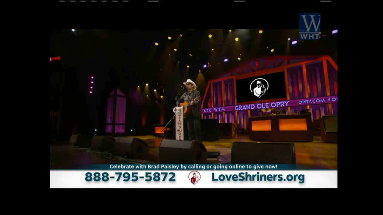 Brad Paisley, in a cowboy hat, strums a guitar center stage at the Grand Ole Opry.  A banner at the bottom promotes a phone number and website for LoveShriners.org.

