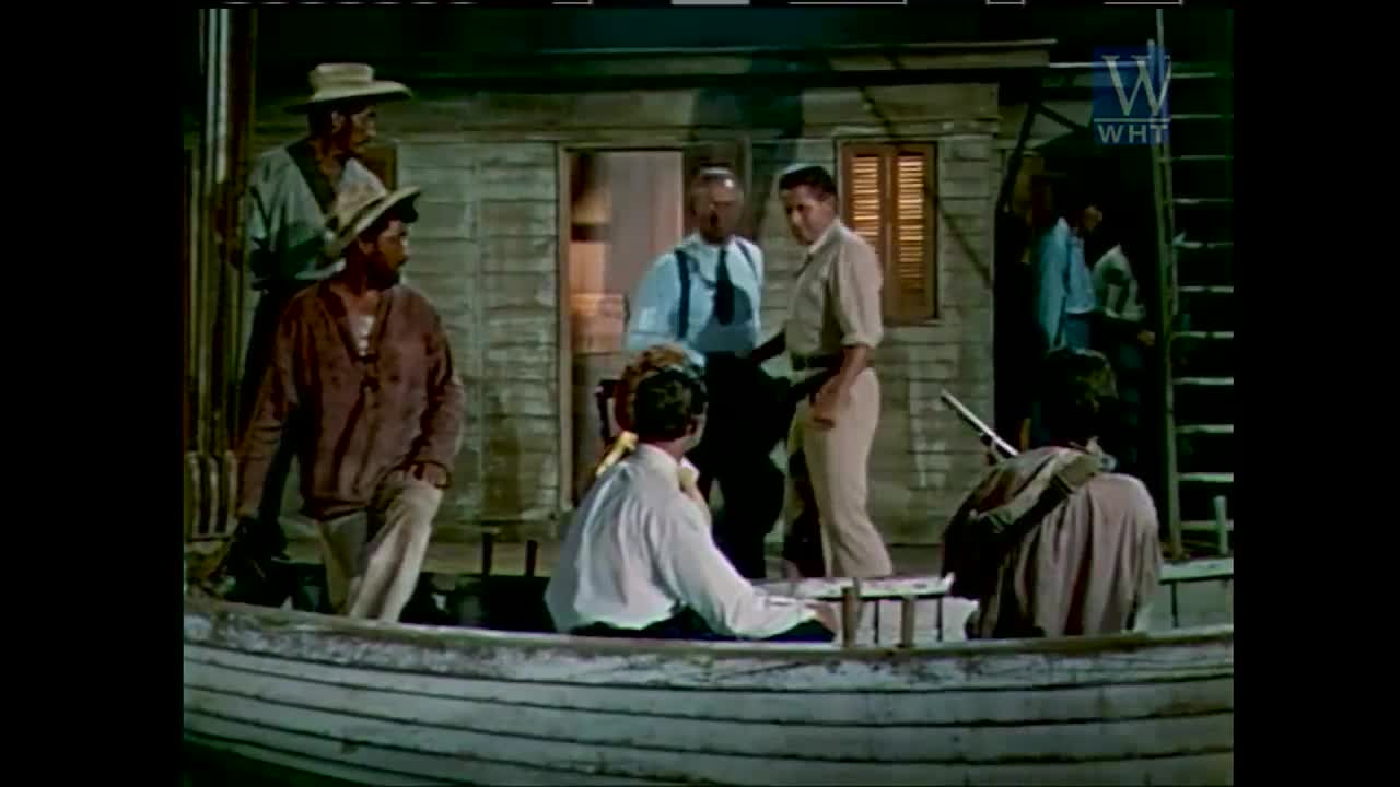 A group of men in the United States, likely from a World Harvest TV production, are gathered near a small wooden structure, with some standing in a boat. The man in the boat is facing away, while others look on, some appearing to be in discussion.
