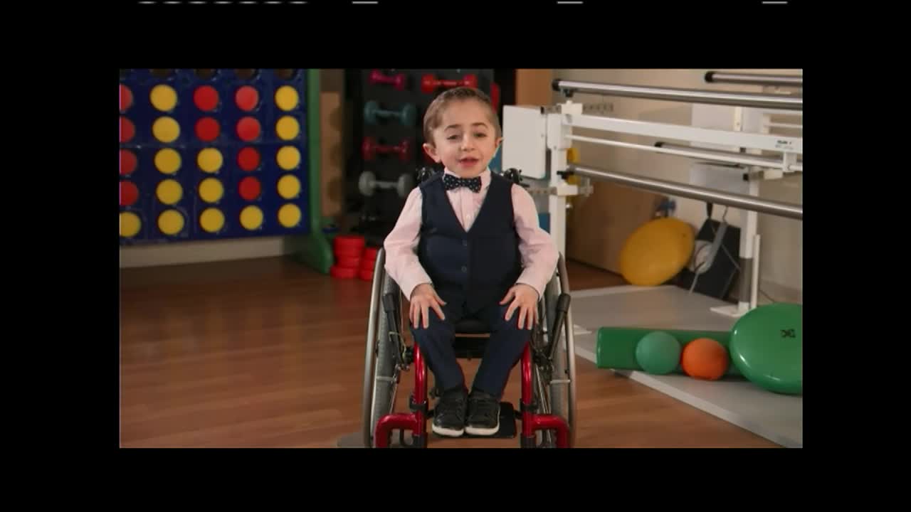 A young boy in a suit and bow tie sits in a wheelchair, speaking directly to the camera. Behind him, exercise equipment and a giant Connect Four game fill the room.
