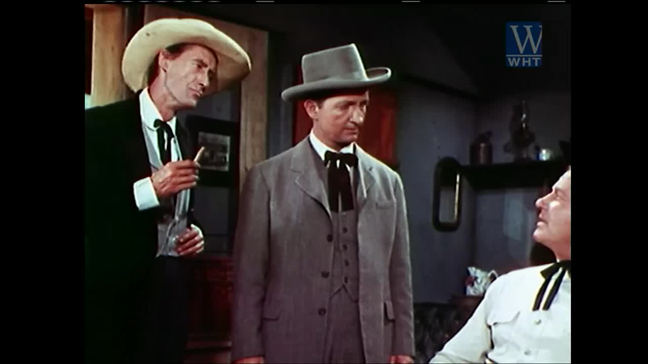 A man in a cowboy hat holds a cigarette and looks to the side, while another in a gray suit stares intently. The third man, seated, looks up at them, his black tie prominent against his white shirt.
