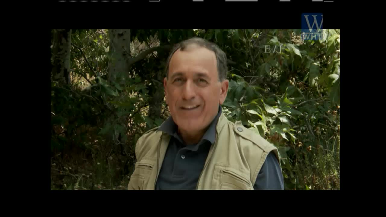 A man in a tan vest smiles, positioned in front of a backdrop of trees. The World Harvest TV logo is visible in the corner.
