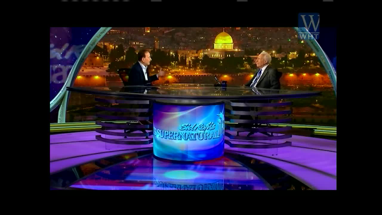 On the set of Sid Roth's "It's Supernatural" on World Harvest TV, two men in suits sit at a large desk. The host gestures with his hands as he speaks to the older man, who listens intently.
