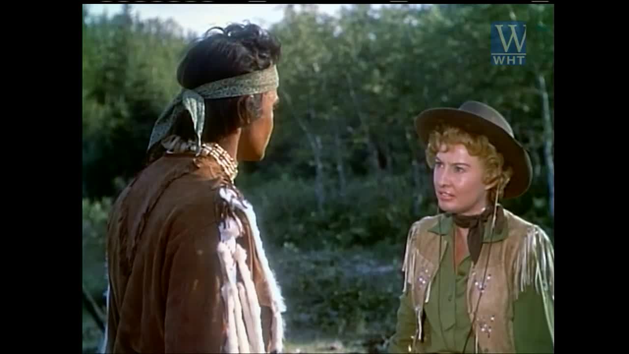 A man in a beaded necklace and headband is turned away from the camera, talking to a woman in a hat and fringed vest. The scene unfolds on World Harvest TV, somewhere in the United States.
