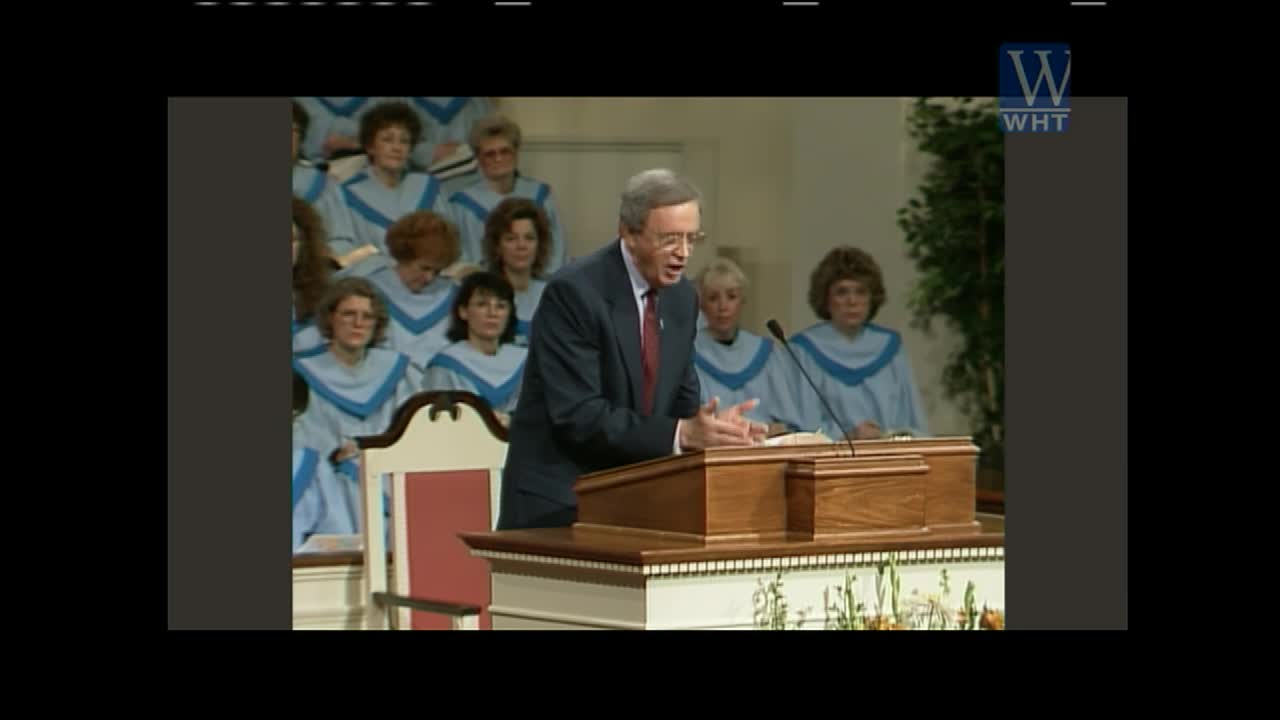 A man in a suit gestures with his hands as he speaks from behind a wooden podium. Behind him, a choir in matching blue robes listens intently.
