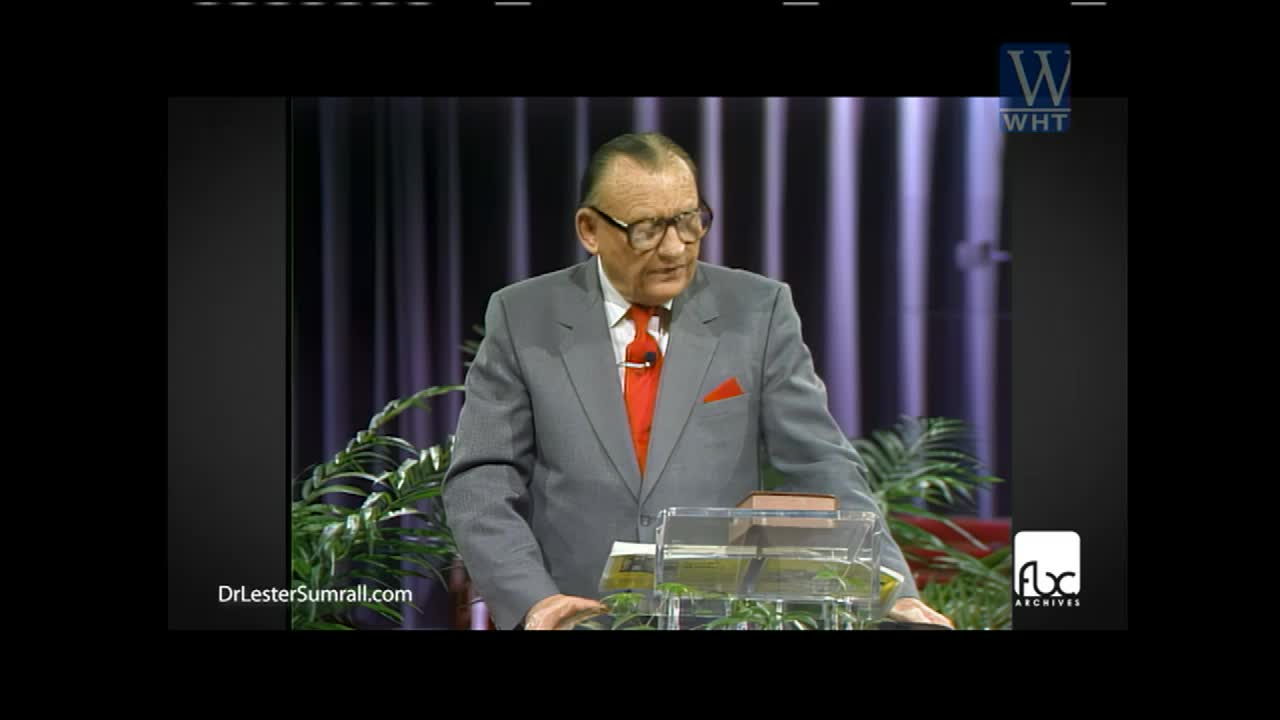A man in a gray suit and red tie stands behind a lectern, speaking on World Harvest TV. He's holding a small book, and there are plants in front of him.
