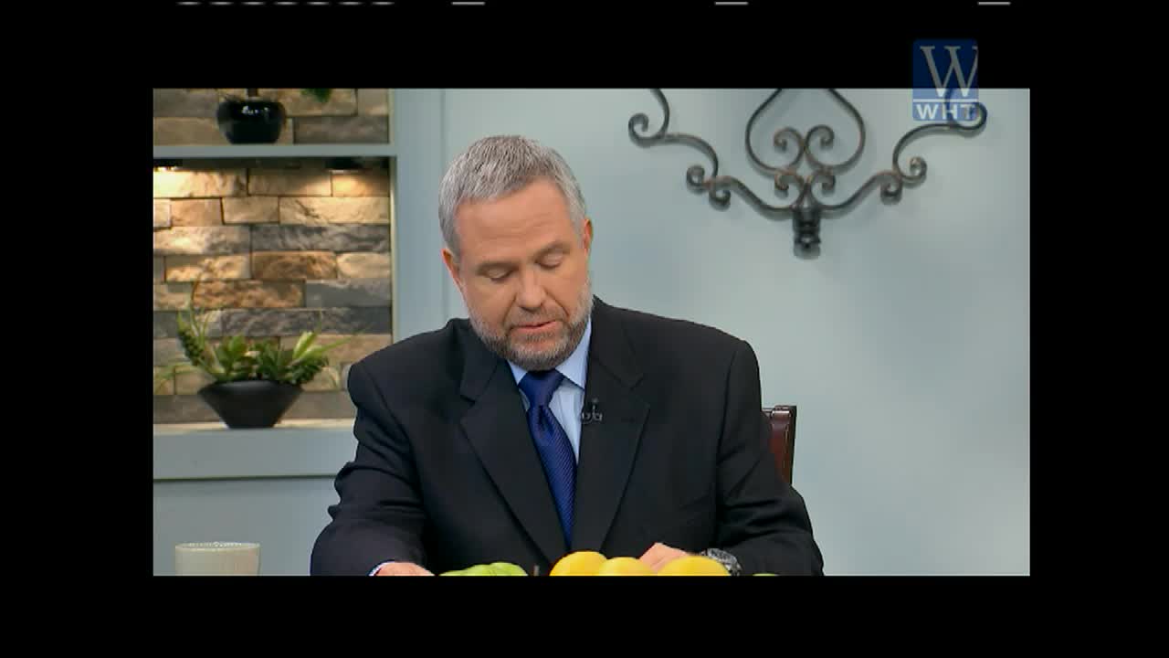 A man in a dark suit, with a blue tie, reads from a script on a table, presumably for World Harvest TV.  Behind him, a decorative wall and a logo for WHT are visible.
