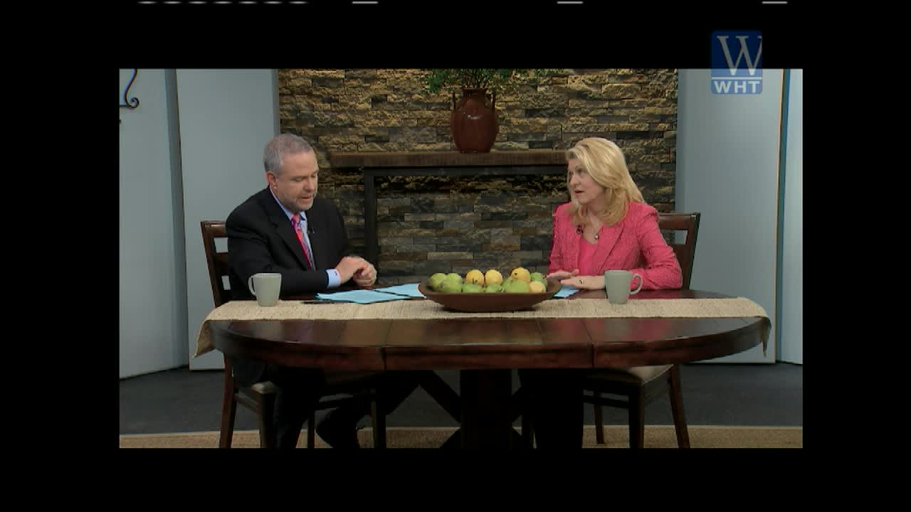 A man in a suit and a woman in a pink jacket sit across from each other at a table, both with mugs and papers in front of them. They're on a set for World Harvest TV, with a stone wall backdrop visible.
