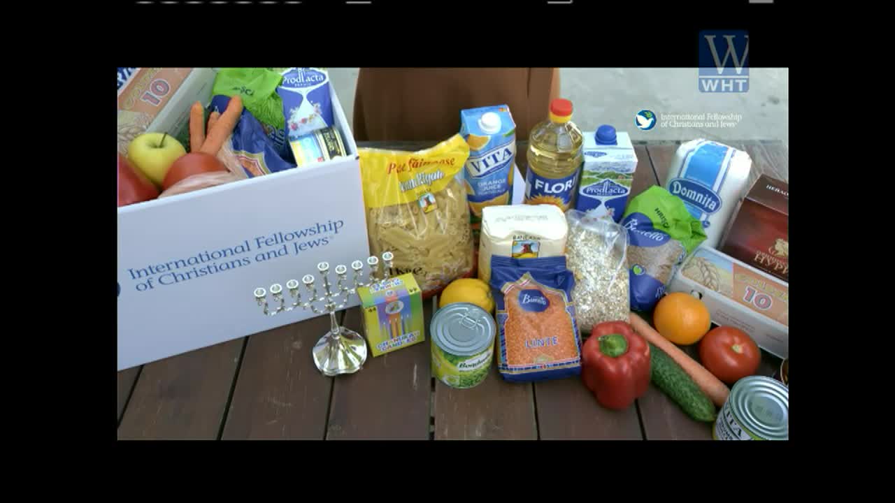 A box overflowing with food, including fresh produce and packaged goods, sits on a wooden table. The International Fellowship of Christians and Jews logo is visible, and the World Harvest TV logo is in the corner.
