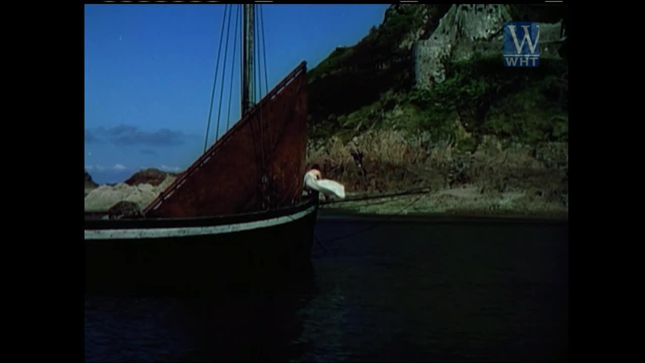 A sailboat with a red sail glides across the water, the dark hull contrasting against the bright blue sky. The scene is presented on World Harvest TV, with a rocky shore and a green hillside in the background.

