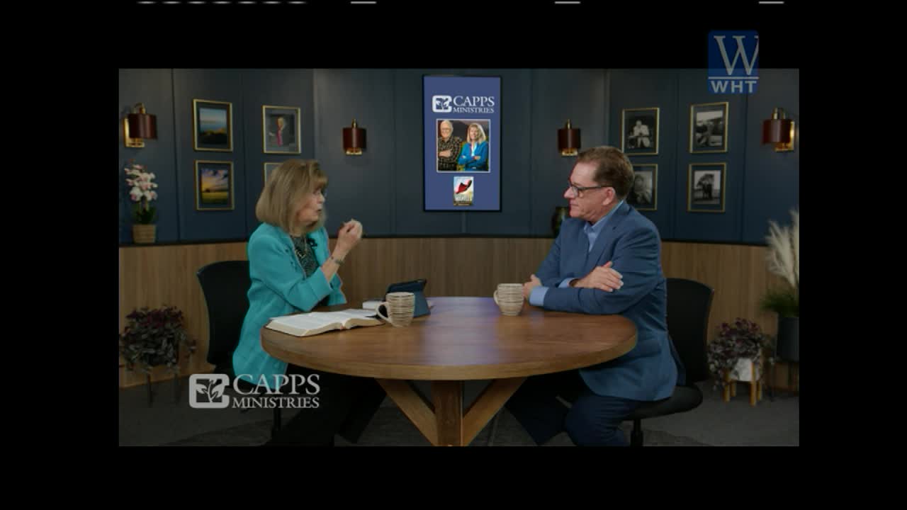 A woman in a teal jacket gestures while speaking to a man in a blue suit across a round table. They are seated in a studio setting, with framed pictures on the wall behind them, likely for a World Harvest Television broadcast.
