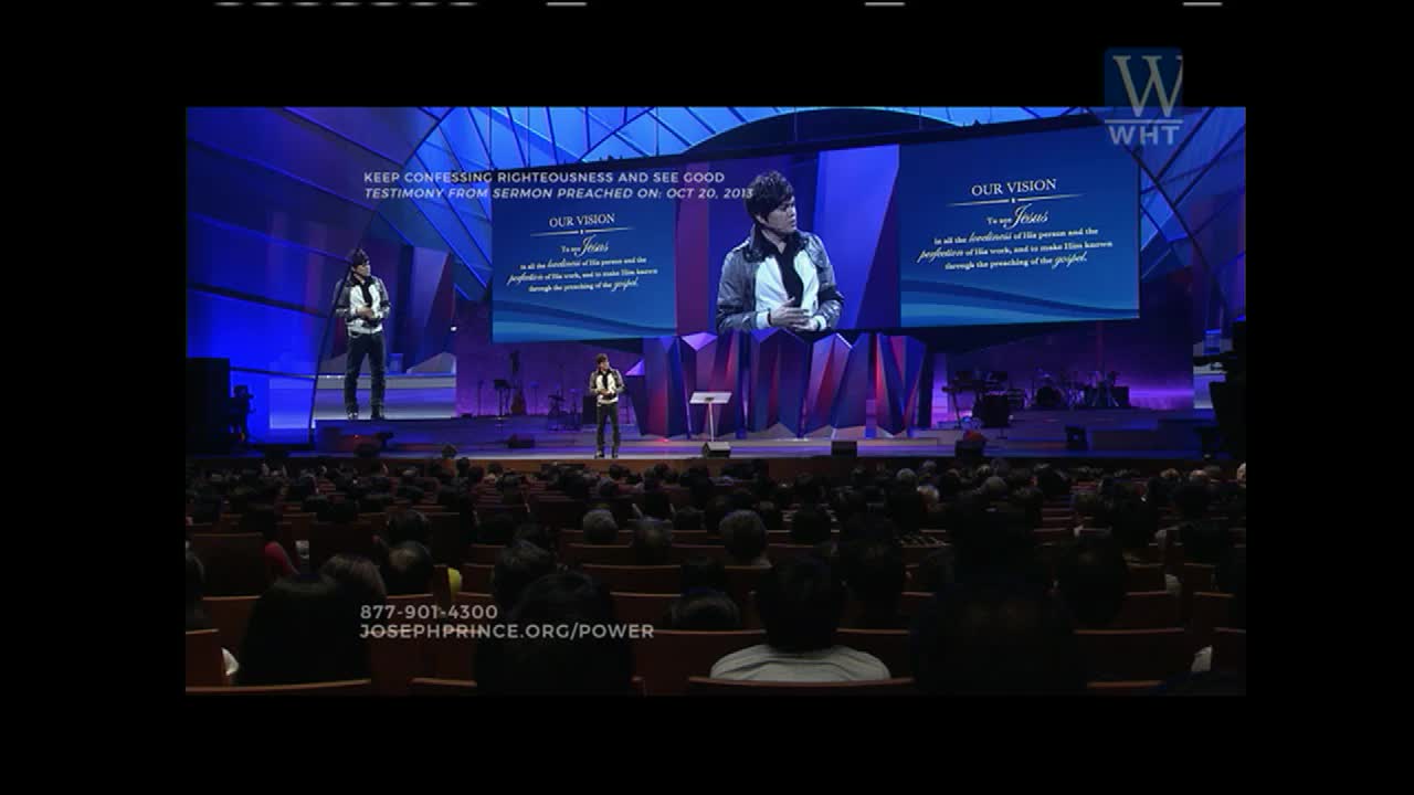 A man in a jacket stands on a stage, speaking to a large audience. Behind him, a screen displays text and a larger image of him.