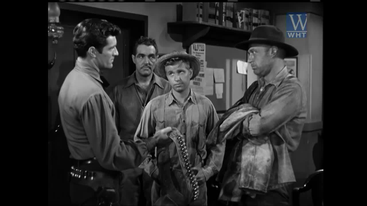 A man in a hat gestures with a belt of bullets to three other men in a dimly lit room. A "Wanted" poster hangs on the wall behind them.