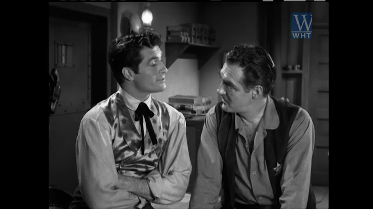 Two men in period Western attire converse intently. The man on the left wears a patterned vest over a collared shirt and a black bolo tie. The man on the right, a sheriff, has a star pinned to his vest.