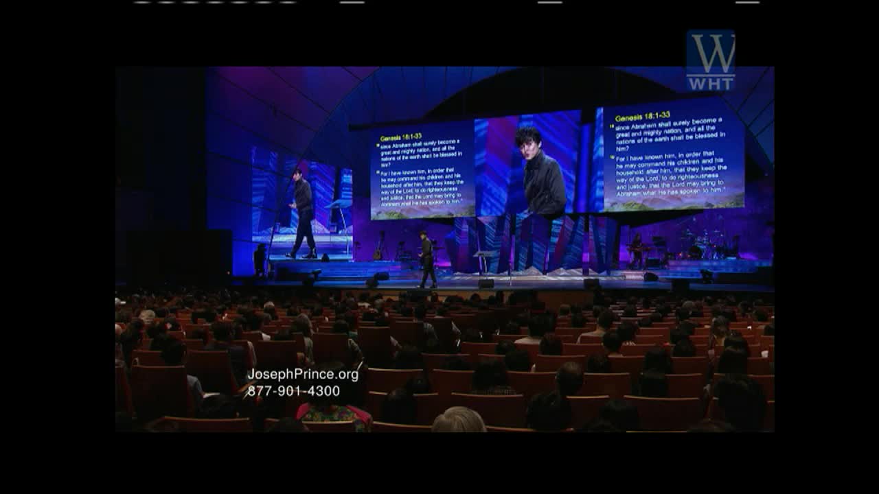 A man in a dark jacket walks across a stage, bathed in blue light. Behind him, two large screens display scripture from Genesis, with his image projected on the right. A packed auditorium watches on.