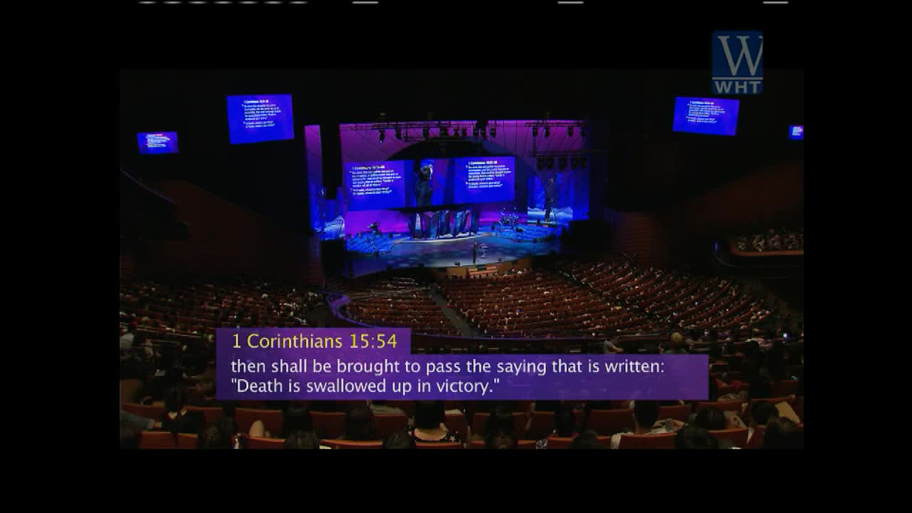 A speaker stands on a stage in a large auditorium, bathed in purple and blue light. The World Harvest TV logo appears in the upper right corner.