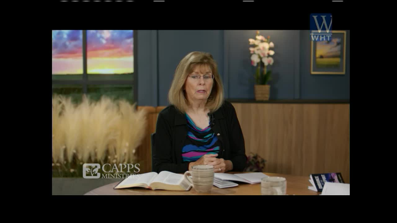 A woman with blonde hair and glasses is speaking at a table. An open Bible and a mug sit before her.