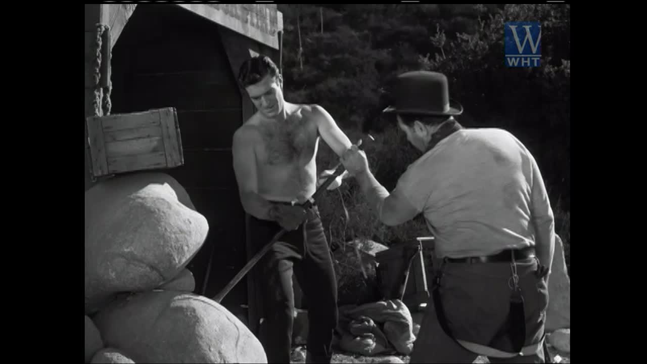 A man, shirtless and muscular, grips a shovel while another man in a bowler hat reaches out to take it. The scene unfolds outdoors, with large rocks and a wooden structure nearby.