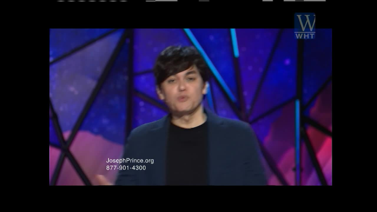 A man in a dark jacket and t-shirt stands on a stage, speaking with his hands gesturing. The backdrop behind him is a geometric pattern of blues and purples.