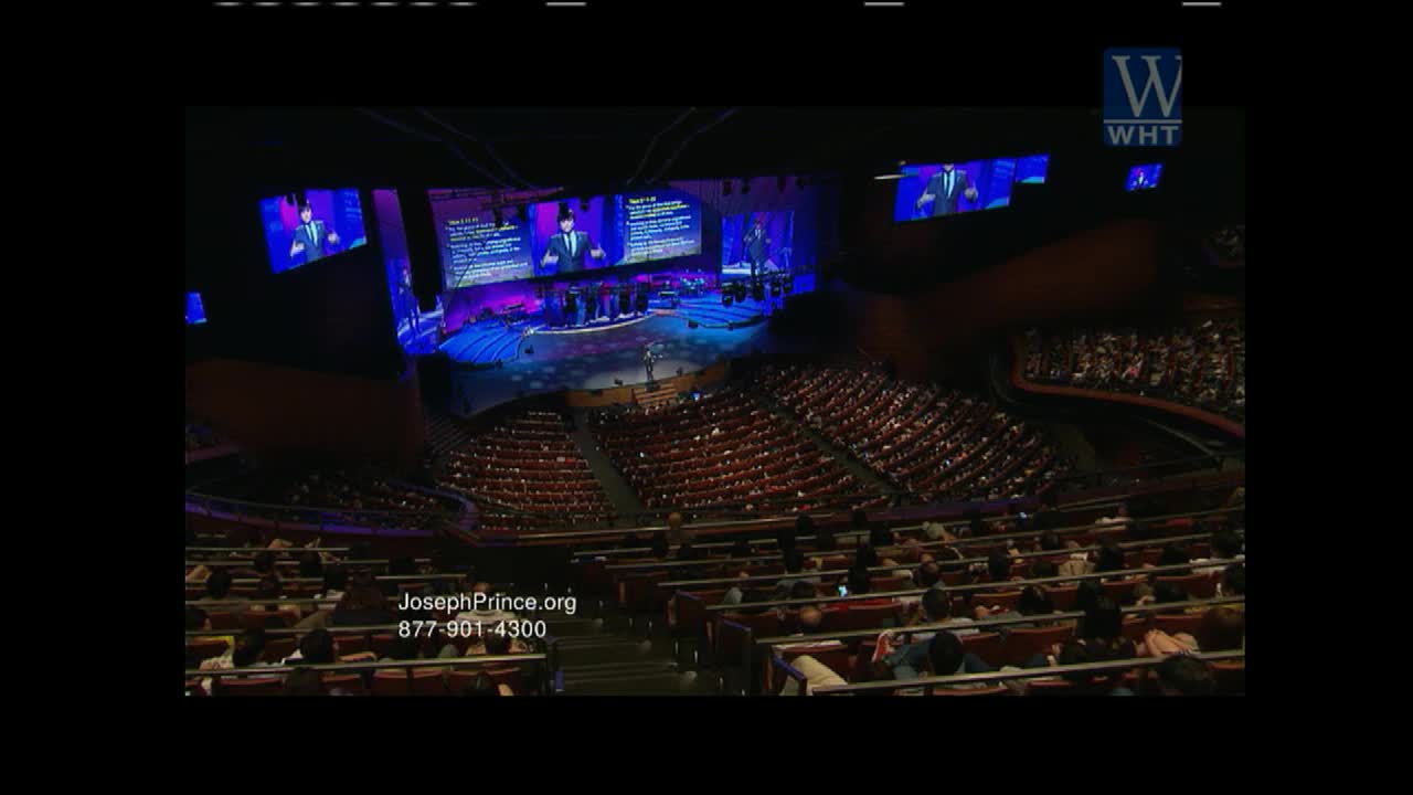 A speaker stands on a brightly lit stage, addressing a large congregation seated in tiered rows. Screens behind him display his image and text, broadcast live on World Harvest TV.