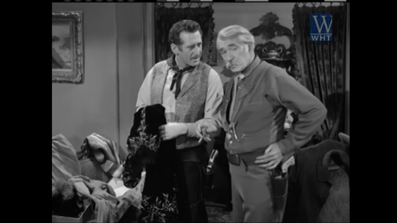 One man holds up a dark, embroidered garment while the other looks on, a cigarette dangling from his lips. The World Harvest Television logo appears in the upper right corner.