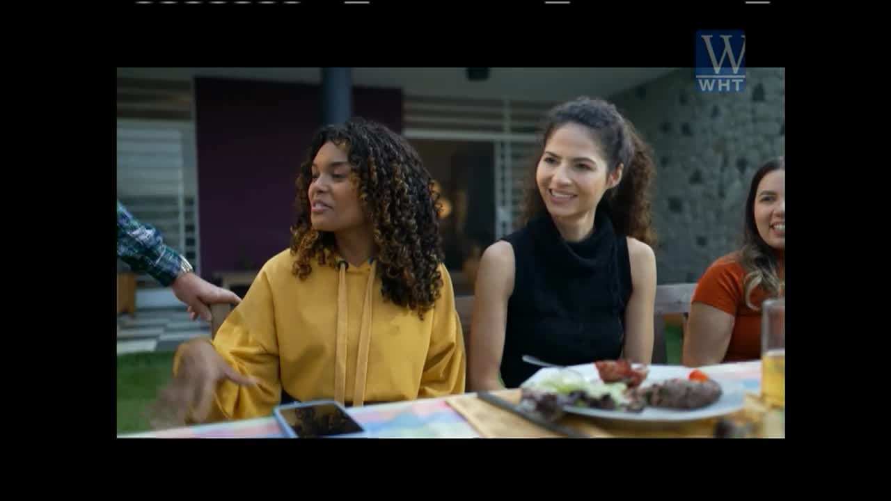 A hand in a plaid sleeve rests on a woman's shoulder as she talks. Another woman smiles beside her at a table laden with food.