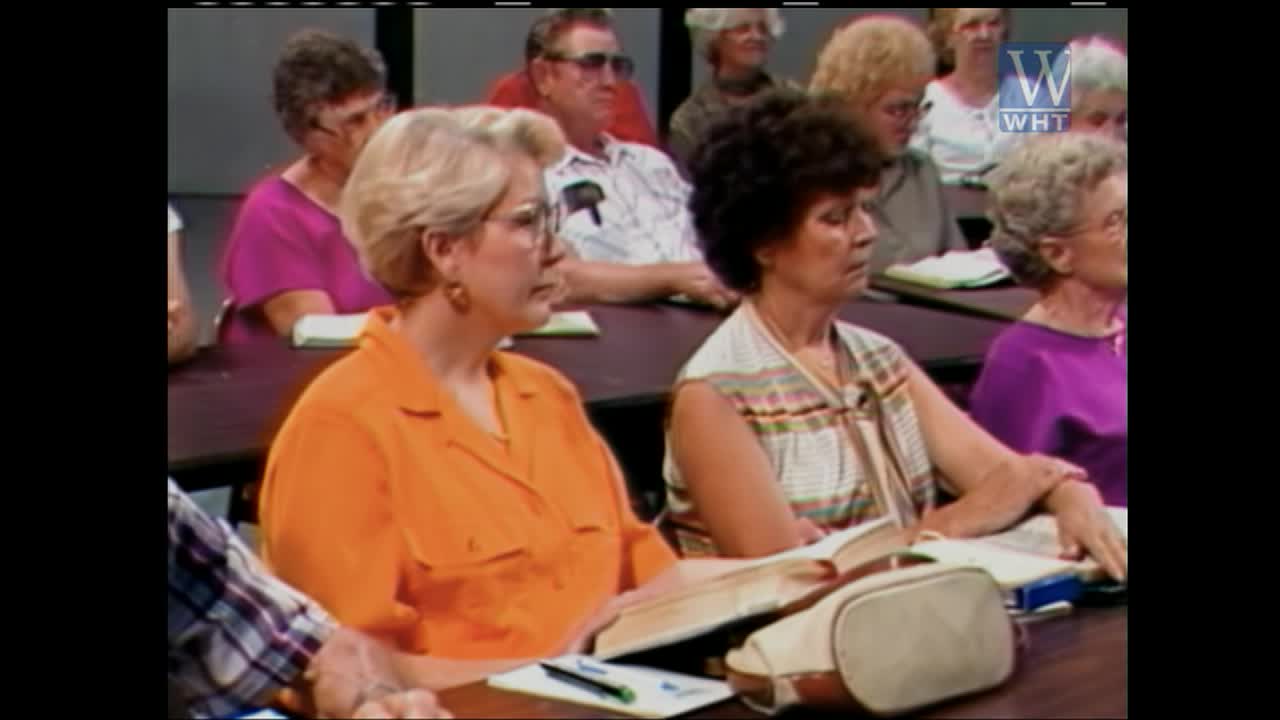 A group of people sit at tables, listening intently. A woman in an orange blouse and another in a patterned top have books open before them.