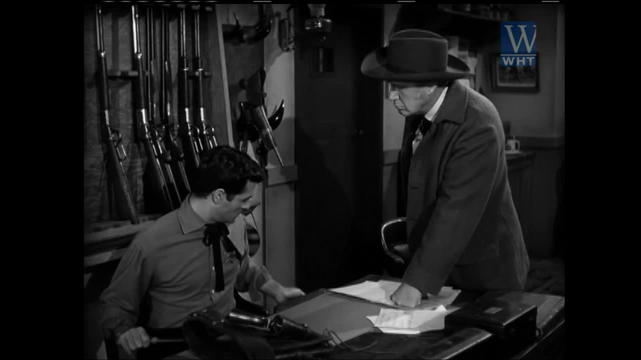 A man in a cowboy shirt leans over a desk, while a man in a suit and hat points at papers. Rifles hang on the wall behind them.
