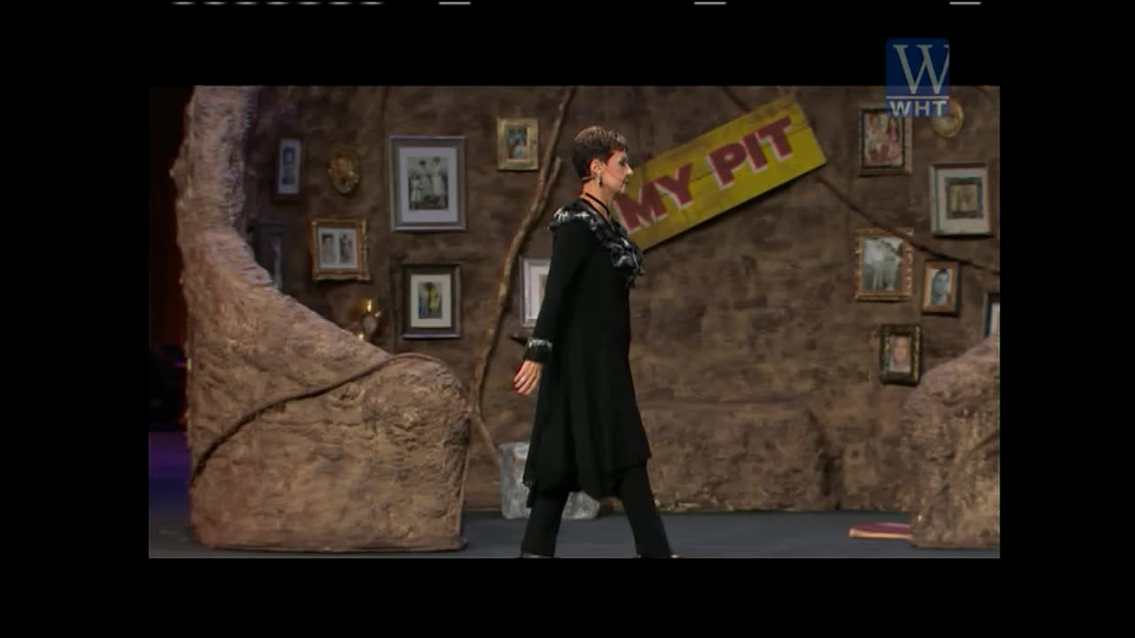A woman in a long black coat walks across a stage set to look like a cave. A yellow sign with "MY PIT" in red letters hangs on the wall behind her.