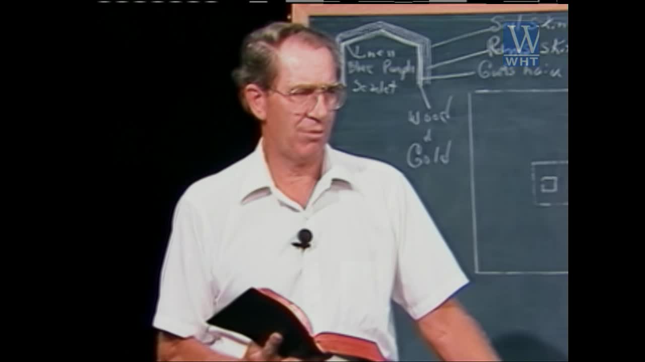 A man in a white shirt stands before a chalkboard, holding an open Bible. He gestures with his left hand as he speaks, while his right hand rests on the book.