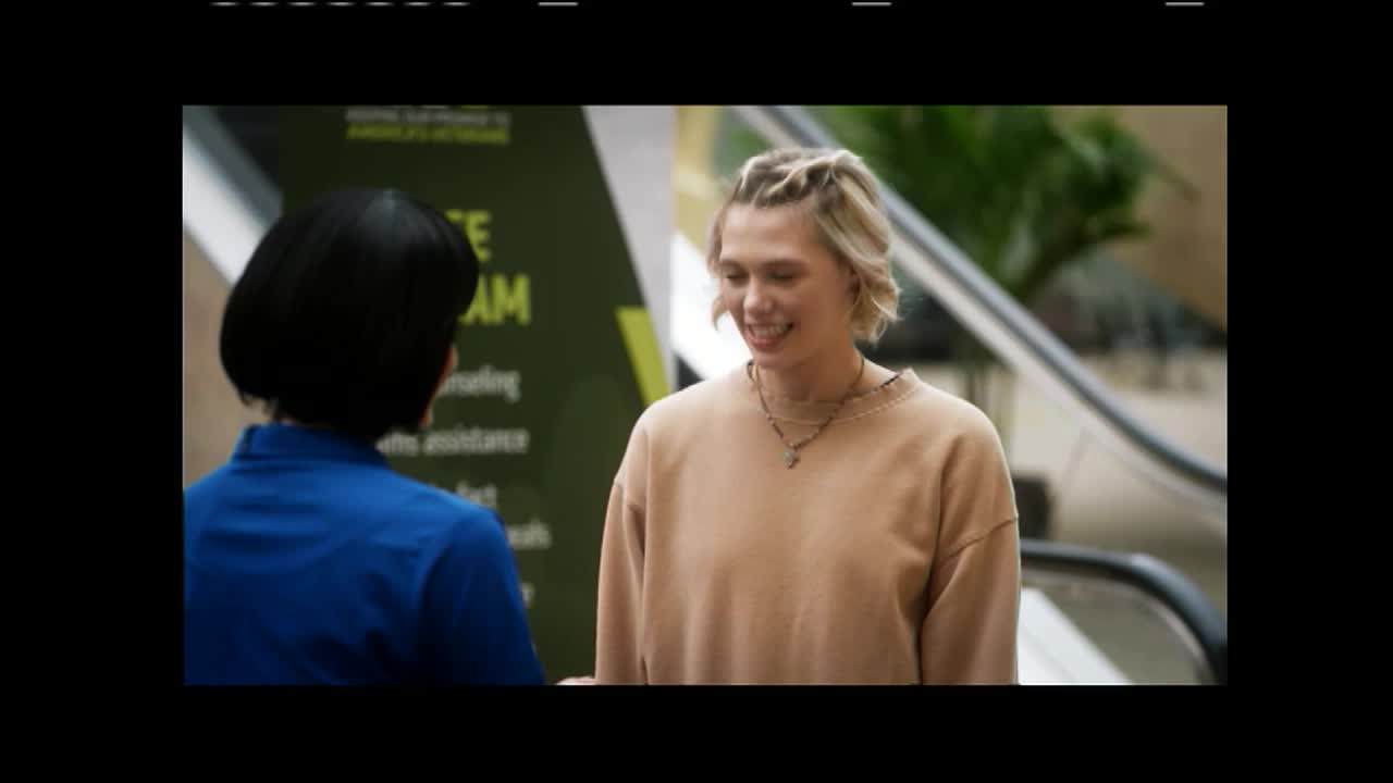 A woman with short blonde hair smiles as she talks to another woman. They stand near an escalator, with a green banner behind them that mentions a program.