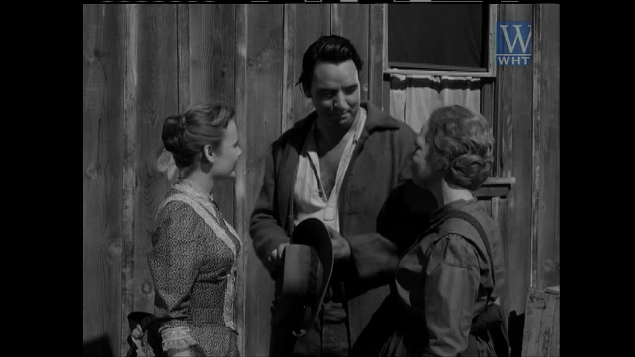 A man stands between two women, holding a hat. The women face each other, their expressions suggesting a shared moment.