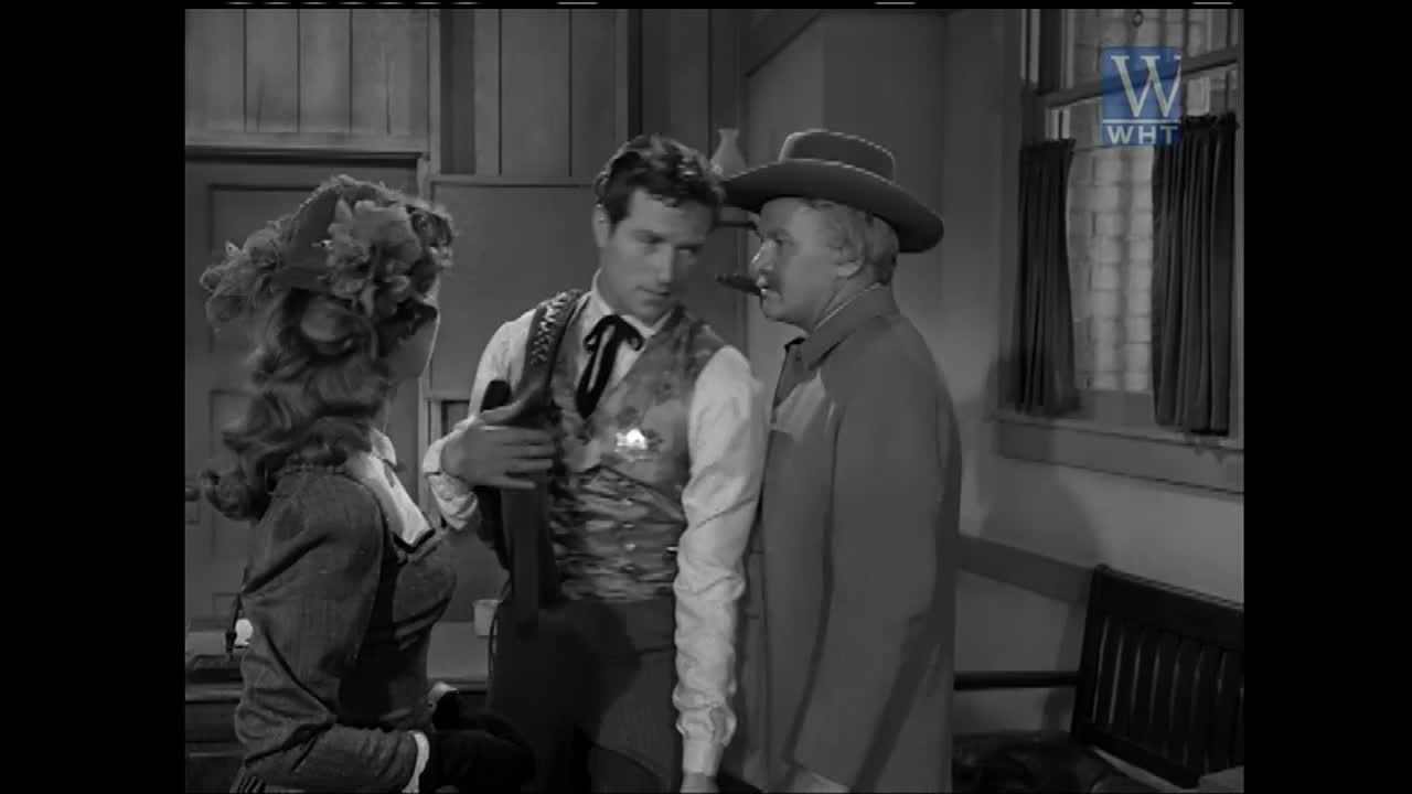 A woman in a wide-brimmed hat and a man in a vest and tie are talking. The man is holding a rifle. Another man, wearing a hat and a long coat, stands nearby, looking at the man with the rifle.