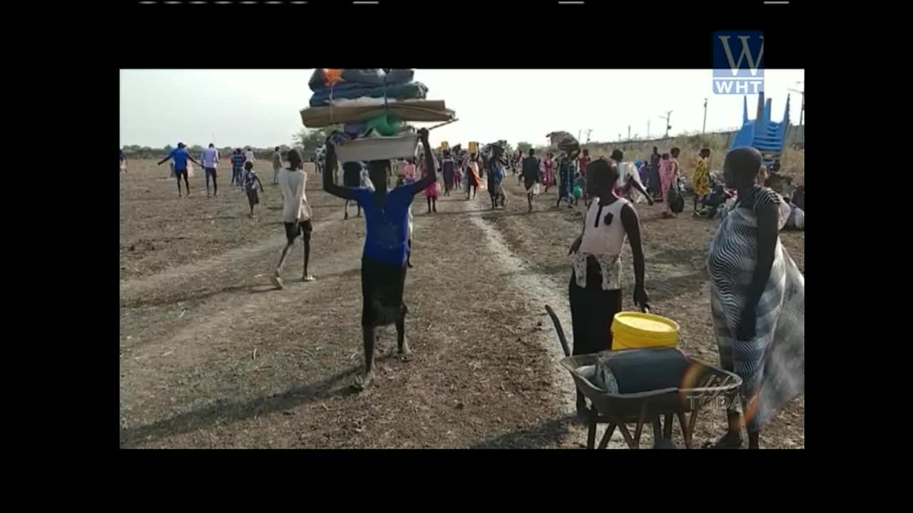 A young person balances a large stack of belongings on their head while walking across a dusty field. Behind them, a procession of people carries their own possessions, some with carts, heading towards a distant blue structure.